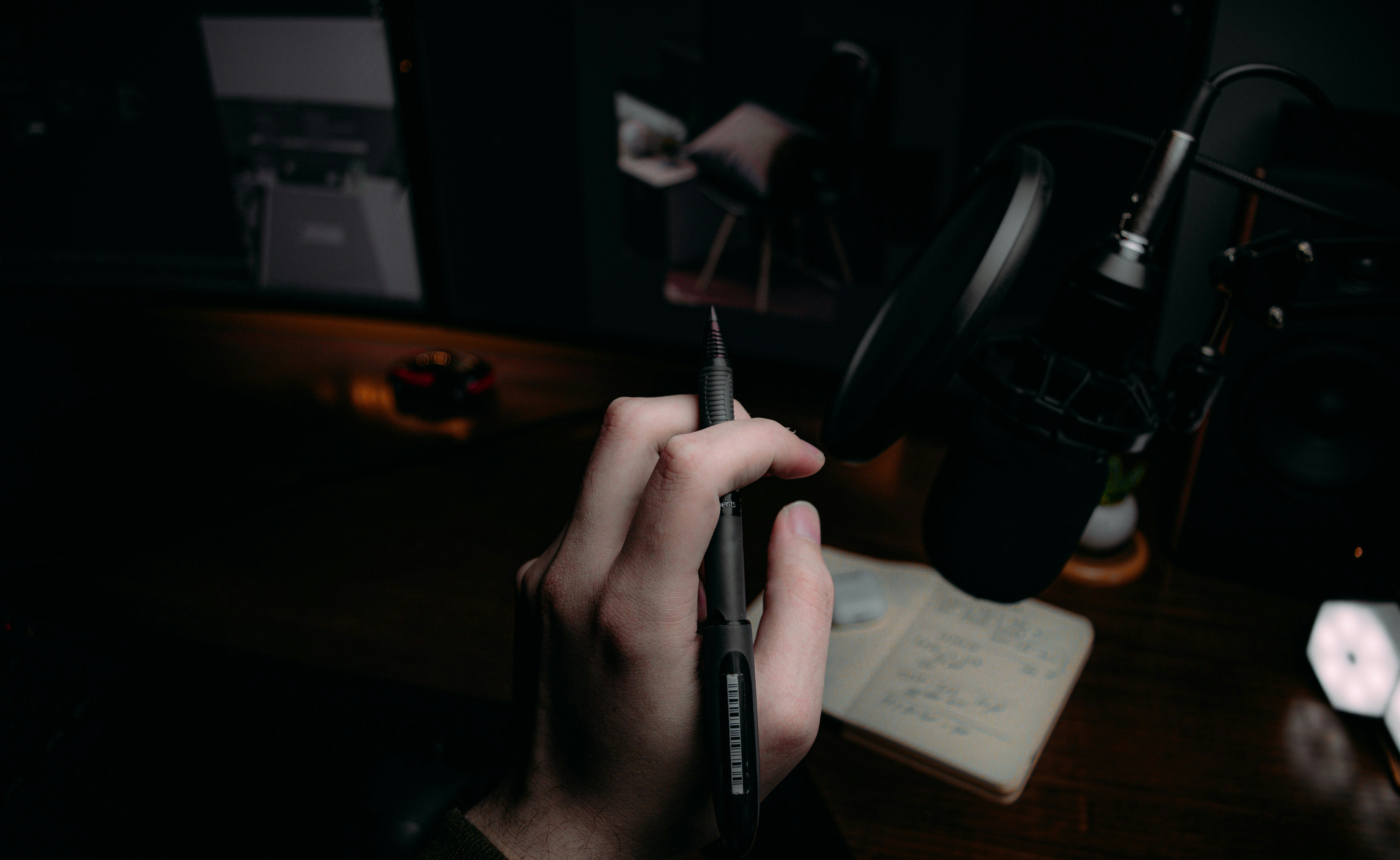 Man holds a pen under a desk lamp near a monitor, meticulously preparing a design