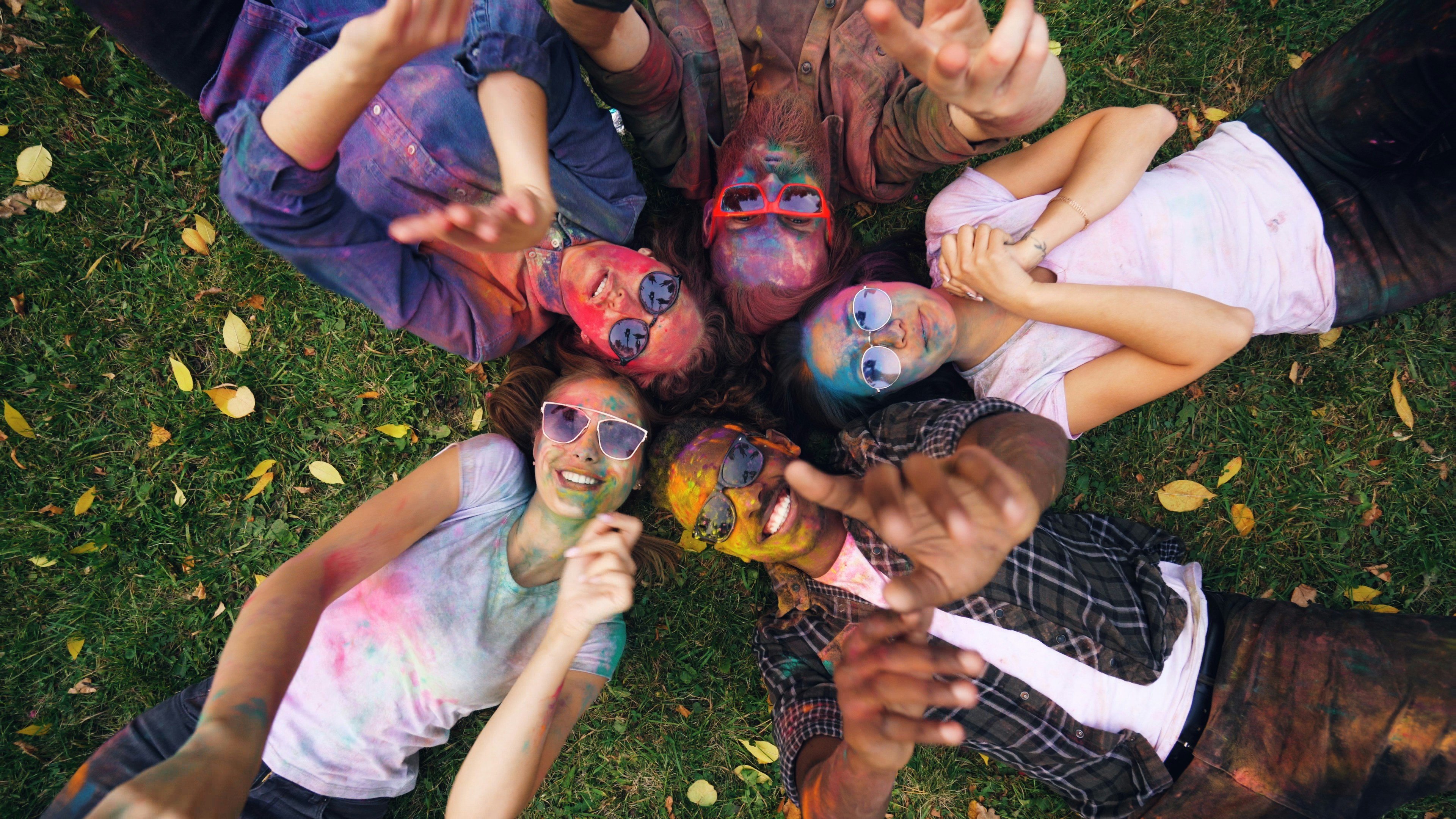 Joyful people, paint-splattered, lie on grass, smiling